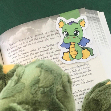 Das Produktbild zeigt ein Magnetlesezeichen, welches in einem aufgeschlagenen Buch steckt. Auf dem Lesezeichen ist ein Drache in den Farben Grün und Gelb. Die Flügel sind blau. Im unteren linken Bildbereich sitzt der dazu passende Plüschdrache.